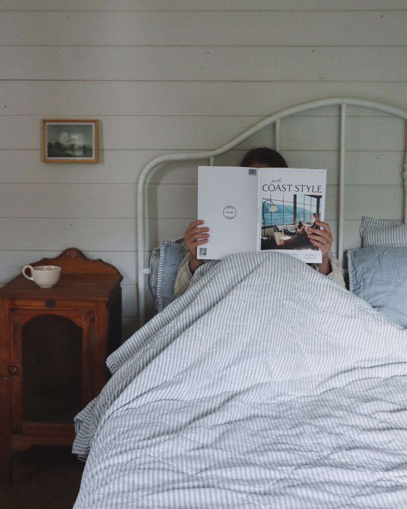 Person lying in bed holding a book titled 'Coast Style' in a bedroom with wooden furniture and a framed picture on the wall.