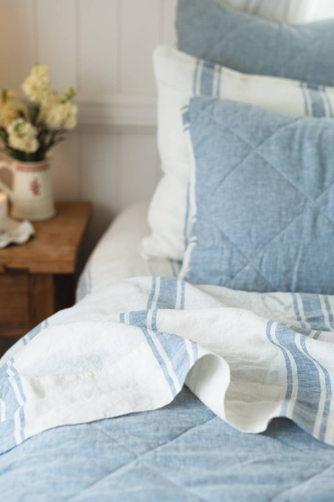 Blue and white quilted bedding on a bed with a side table and flowers in the background.