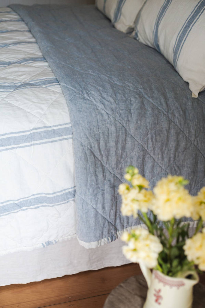 Blue quilt on a bed with white and blue striped bedding, and a vase of yellow flowers in the foreground.