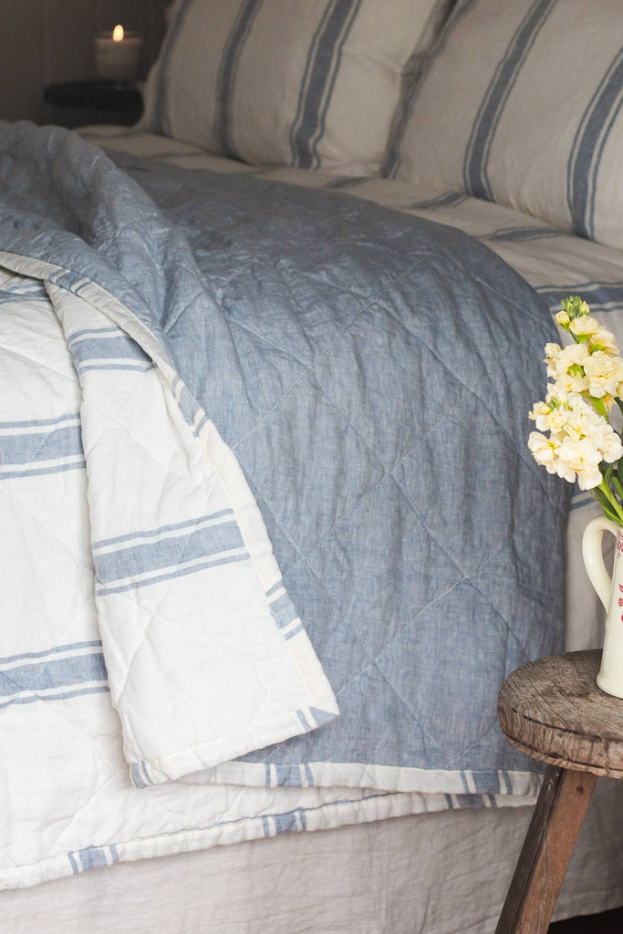 Striped bedding with a blue throw blanket on a bed, next to a small wooden table with flowers.