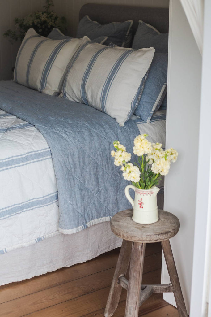 Bed with striped bedding and pillows, accompanied by a small wooden stool with a vase of flowers.