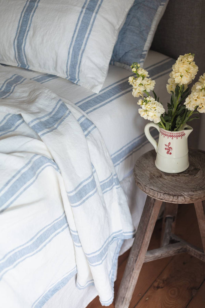 Bed with striped bedding and pillows, wooden stool with flowers, and floral-patterned pitcher.