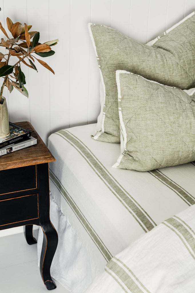 Bed with striped bedding and green pillows next to a wooden nightstand with a plant.