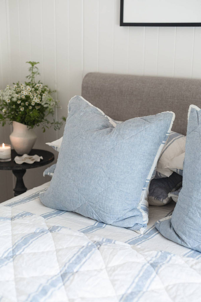 Bed with blue pillows and a gray headboard, featuring a small table with flowers and a candle.