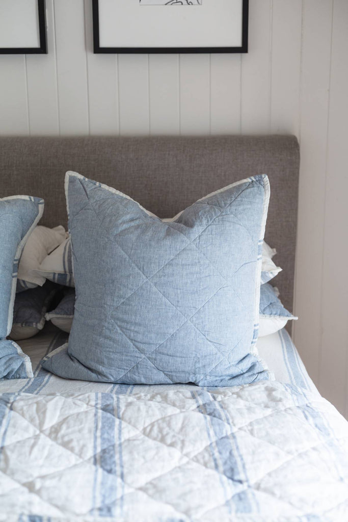 Blue quilted pillow on a bed with a neutral headboard and wall.